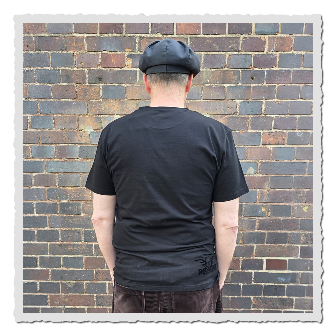 Rückansicht einer Person im schwarzen T-Shirt vor Backsteinwand, kleines dunkles Logo unten rechts sichtbar.